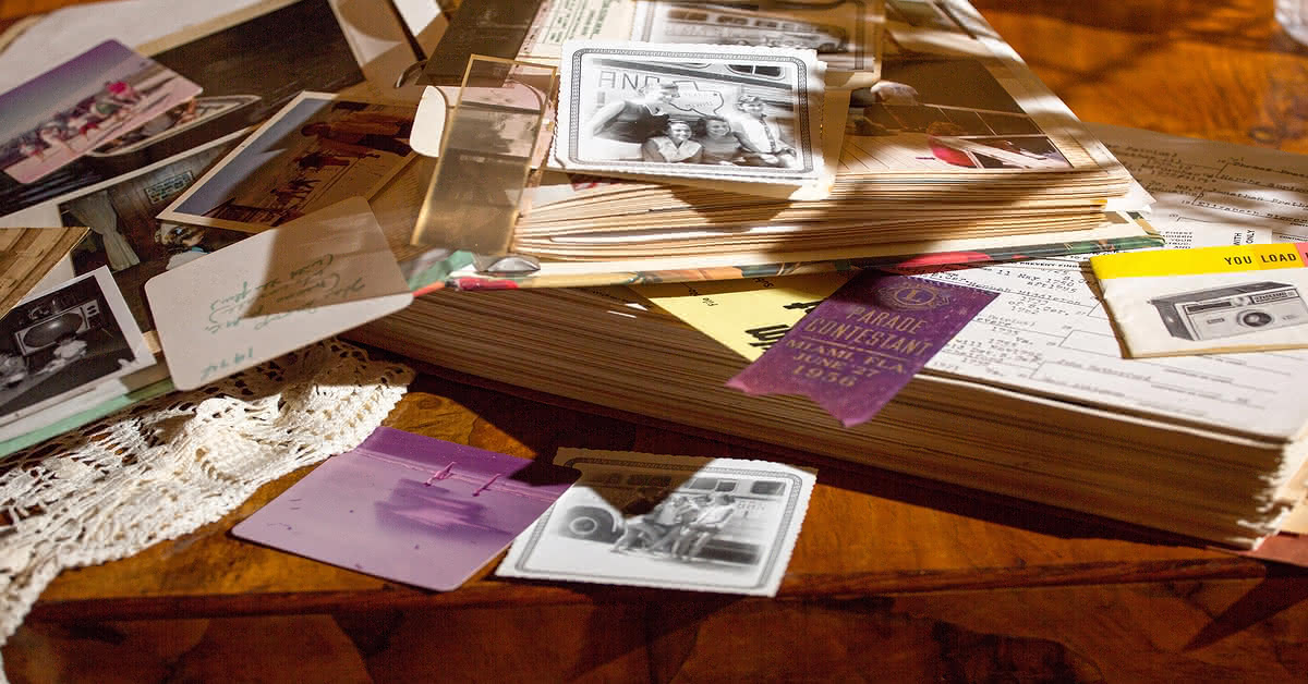 A scrapbook lying open on a wooden table, old photographs and various paper scraps and memorabilia spread over the open pages and parts of the table.