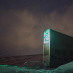 A person with a headtorch approaching the entrance to the Svalbard Global Seed Vault, at night. Ground lit with green light from the top of the entrance, sky with a purple hue, showing stars.